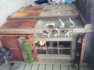 A small rustic shed with wood siding, metal accents, and plants. There are bells and decorations on the walls.