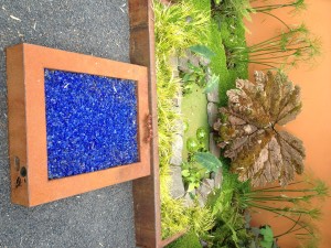 A garden with a large leaf sculpture, small pond, and a rectangular fire pit filled with blue stones.