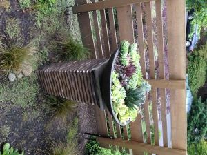 A dark planter, filled with succulents, sits on a textured, brown pedestal, in front of a wooden fence.