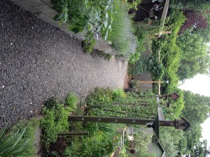 A gravel path winds through a garden with raised beds, bushes, and birdhouses. 