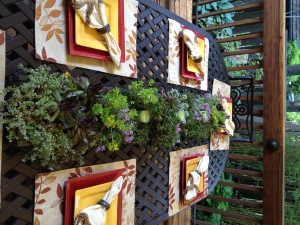 A dark metal outdoor table with a long arrangement of greenery and small plants running down the center, set with red and yellow plates, napkins, and placemats.