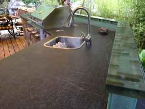 Dark gray countertop with a stainless steel sink and faucet, part of an outdoor kitchen. 