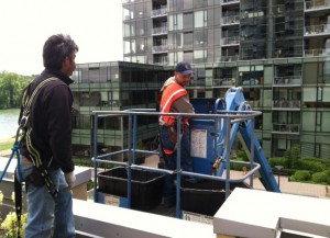 Two men in safety gear, one on a tall blue lift platform, are working on a building's exterior. 